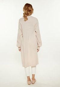 Cardigan lungo di un pallido rosa con una vestibilità comoda, polsini a coste e una superficie liscia. Indossato sopra pantaloni bianchi e scarpe slip-on di colore chiaro.