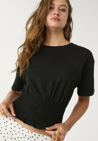 Maglietta nera a maniche corte con vita arricciata, scollo rotondo e morbido materiale di cotone. Indossata con pantaloni bianchi a fantasia.