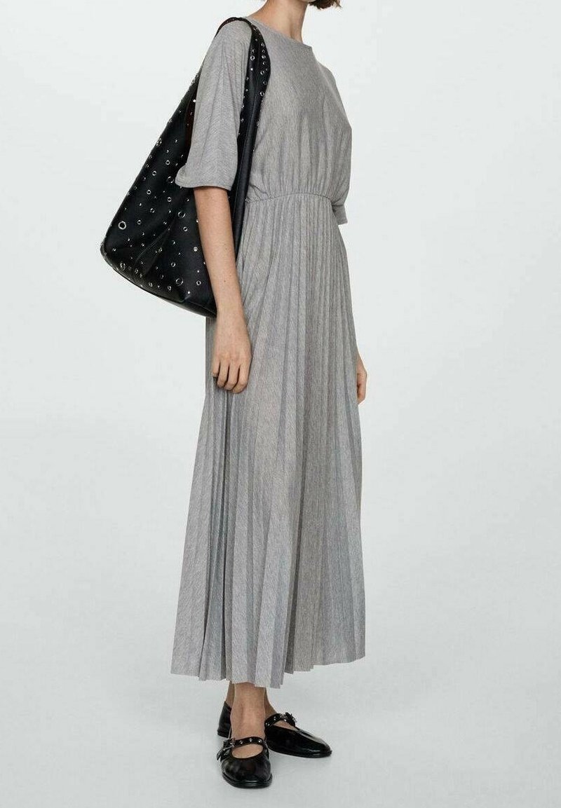 Femme portant une longue robe grise plissée et des chaussures plates noires, portant un grand sac noir à bandoulière avec des œillets métalliques.