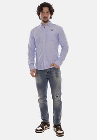 Camisa de botones de manga larga azul claro con un bolsillo en el pecho, combinada con jeans azules desgastados y zapatillas marrones con detalles en blanco.