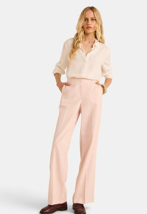 Vrouw met lang blond haar, gekleed in een crèmekleurig blouse en lichtroze, wijde broek met hoge taille, met bruine schoenen, staand met handen in de zakken.