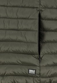 Dunkelgrüner, gesteppten Jackenabschnitt mit horizontaler Steppung, einer teilweise offenen vertikalen Reißverschlusstasche und einem Etikett "Original 86 Always Be Bold Sublevel".