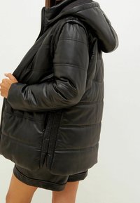 Chaqueta negra acolchada de cuero, con capucha, cierre de cremallera, patrón acolchado y bolsillos laterales. Textura suave y corte relajado.