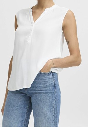 Osoba nosząca białą, bezrękawną, zapinaną na guziki bluzkę włożoną do jasnoniebieskich dżinsów, z jedną ręką w kieszeni i widocznym małym tatuażem na ramieniu.