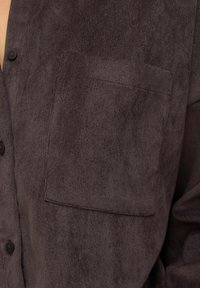 Bruine corduroy shirt met een knoopsluiting en een enkele borstzak. De textuur van de stof heeft een zachte, geribde afwerking.