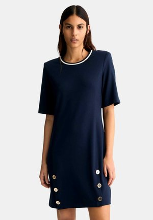 Abito corto blu navy con maniche corte, caratterizzato da un colletto tondo bianco e bottoni decorativi dorati lungo il lato sinistro. Tessuto dalla consistenza liscia.