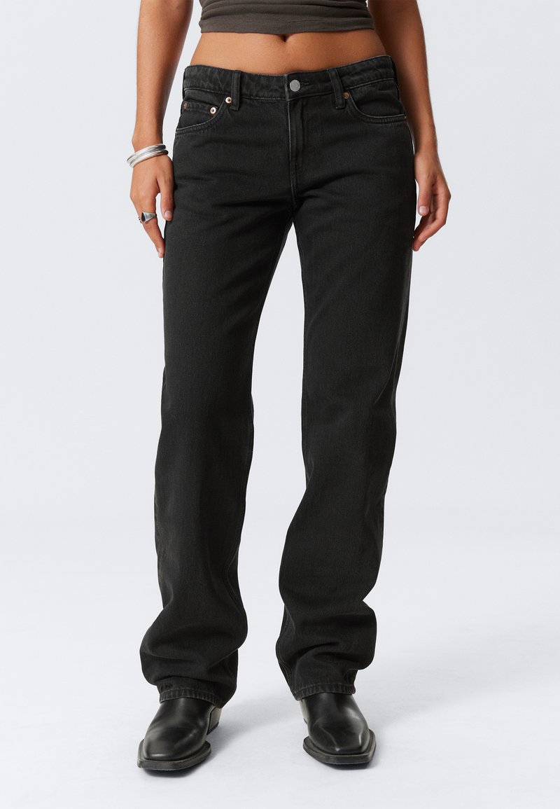 Naine kannab kõrge vöökohaga musti teksaseid ja musti saapaid, seisab käed rahulikult külgedel, paljastades keskmise osa kõhust ja hõbedased käevõrud vasakus randmes.