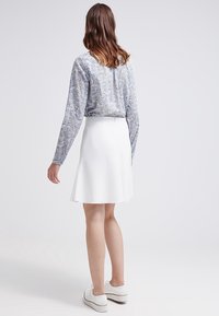 Weißer A-Linien-Rock kombiniert mit einer blauen gemusterten Bluse mit langen Ärmeln, die einen runden Ausschnitt und strukturierten Stoff aufweist. Lässiges Outfit mit weißen Schuhen.