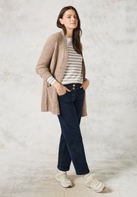 Un cardigan en tricot beige porté sur une chemise à manches longues rayée grise et blanche, associé à un jean bleu foncé et des baskets clair.