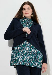 Cardigan navy con motivo testurizzato indossato sopra una blusa floreale teal e bianca. L'outfit presenta una vestibilità rilassata e materiali morbidi.