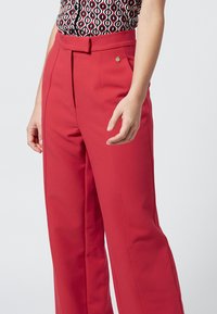 Red wide-leg trousers with a smooth texture, high waist, and front pleats. Features a gold button accent and styled with a patterned top.