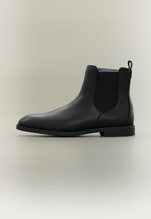 Botte Chelsea en cuir noir avec panneaux latéraux élastiques et languette, talon bas et bout arrondi lisse sur fond neutre.