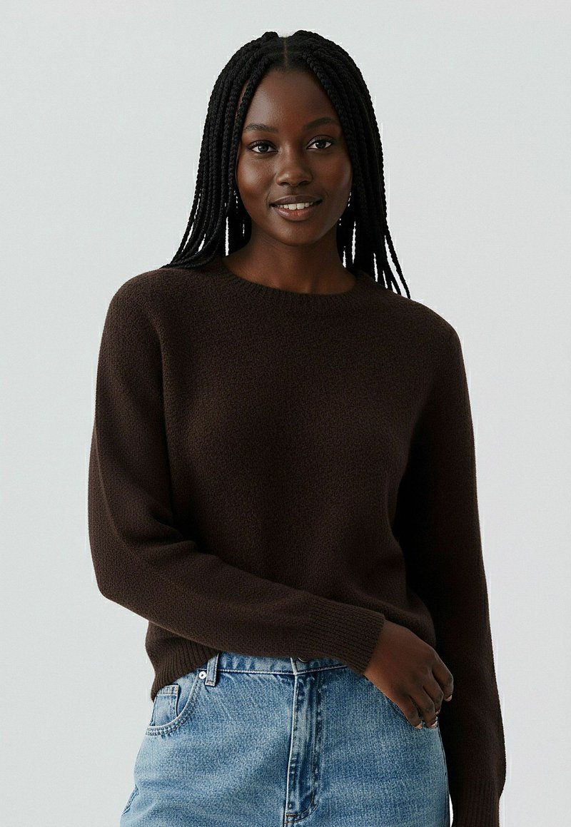 Pull en maille marron avec col rond et manches longues, associé à un jean bleu clair. Le tissu semble doux et chaud.