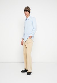 Camisa azul clara às riscas com botões, calças bege e mocassins pretos. Gorro de malha às riscas castanhas e pretas. Modelo está em pé com uma mão no bolso.