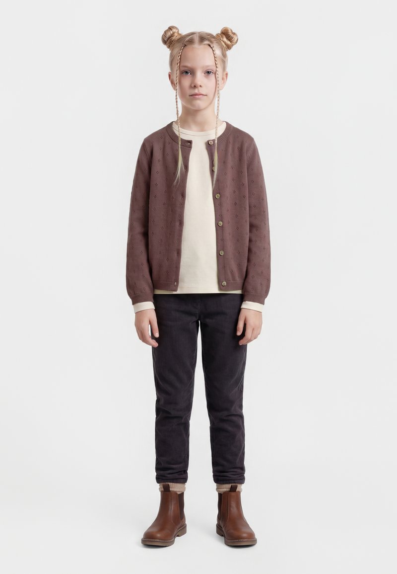 Ung person stående, iført brun knappet cardigan, cremefarvet skjorte, sorte bukser, brune støvler, med håret stylet i to knolde og to frontfletninger.