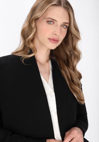 Schwarze Strickjacke mit V-Ausschnitt, weicher Textur und langen Ärmeln, getragen über einer weißen Bluse mit Knopfdetails. Haare in lockeren Wellen gestylt.