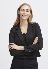 Fekete rövidített blézer, amelynek hajtókája és hosszú ujjai vannak, sima anyagból készült. Egy fekete csipkés részletekkel díszített felső fölött viselik.