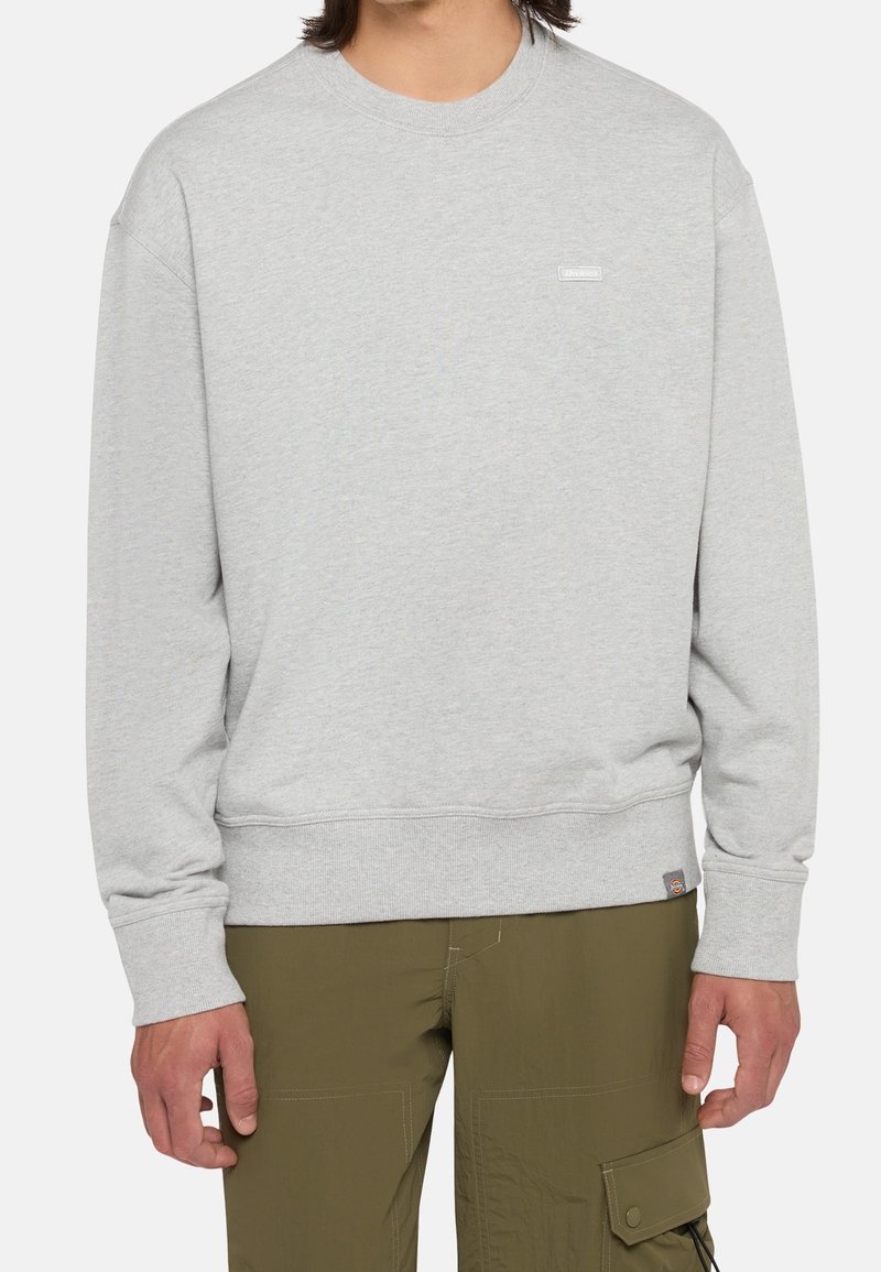 Homme portant un sweat-shirt gris clair à col rond et un pantalon cargo vert olive, debout les bras détendus le long du corps.