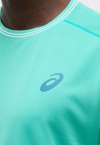 Κοντινό πλάνο ενός ατόμου που φοράει ένα τιρκουάζ αθλητικό μπλουζάκι με ανοιχτό μπλε λογότυπο Asics στο στήθος και λεπτομέρειες γιακά με άσπρες ρίγες.