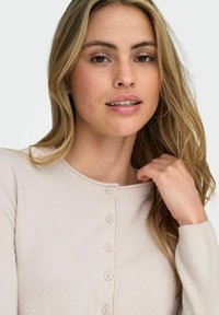 Lys beige cardigan med rund halsudskæring, der har syv runde knapper foran og ribstrikkanter. Blød tekstur og tætsiddende design.