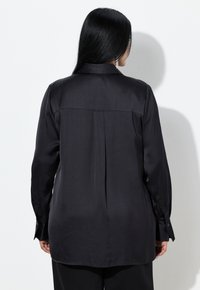 Sort satin skjorte med spids krave, lange ærmer og en central læg på ryggen, med knaplukning ved manchetterne og en glat tekstur.