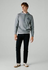 Felpa grigia con tasca frontale e cappuccio con cordino, caratterizzata da un logo sul petto; abbinata a jeans scuri e sneakers bianche.