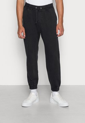 Uomo che indossa pantaloni jogger neri con vita elastica e coulisse e sneakers bianche, in piedi su pavimento grigio e sfondo bianco.