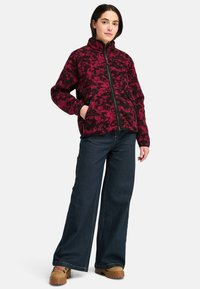 Giacca in pile a motivo rosso e nero con colletto alto e chiusura con zip, abbinata a jeans in denim scuri a gamba larga e calzature marroni.