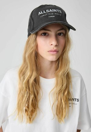 Ung person med langt blondt hår iført en sort AllSaints-kasket og hvid AllSaints t-shirt mod en ensfarvet baggrund.