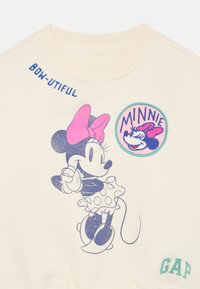 Kremfarget hettegenser med grafikk av en tegneseriefigur i prikkete kjole, tekst "BOW-UTIFUL," og "MINNIE" applikasjon i rosa og teal.
