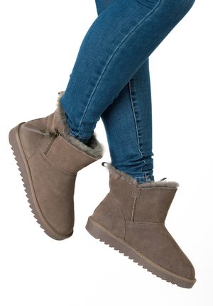 Persoon die middenkuitlange winterlaarzen van bruin suède draagt met een bontvoering en een ritssluiting aan de zijkant, gecombineerd met een blauwe skinny jeans tegen een witte achtergrond.