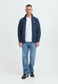 Mladý muž stojí čelom dopredu, oblečený v námorní modrej zipovej bunde cez bielo tričko, v voľných modrých džínsoch a čiernych teniskách s bielymi pruhmi.