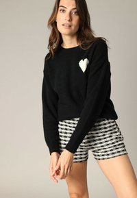 Pull en maille noir avec un accent en forme de cœur blanc ; poignets côtelés. Associé à un short à motifs avec des rayures noires et blanches.
