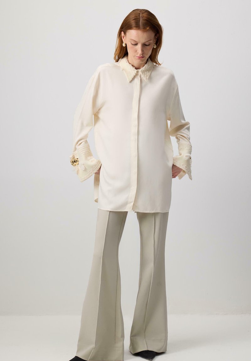 Blouse crème avec un devant à boutons, un col texturé et des manches évasées. Assortie à un pantalon évasé beige clair. Tissu lisse.