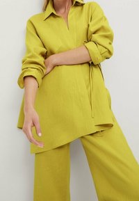 Limoninvihreä pellavapaita, jossa on kaulus ja käärityt hihat, yhdistettynä sopivasti löysiä housuja, joissa on vyötäröllä solmimisnauha muodon vuoksi.