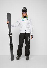 Skiausrüstung mit einer weißen, geknöpften Jacke mit Taschen, schwarzen Hosen und schwarzen Skistiefeln; hält einen schwarzen Ski; bunter Schal und Mütze.