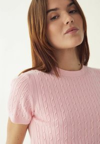 Hele roosa lühikeste varrukatega kootud kampsun, millel on tekstuuriga kaablisilmusmuster ja ümargune kaelus, valmistatud pehmest, hingavast materjalist.