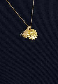 Pendentif soleil en or avec un visage serein et un pompon beige sur une fine chaîne en or sur un tissu foncé texturé.