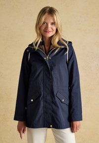Blonde Frau trägt eine marineblaue Kapuzen-Regenjacke mit goldfarbenem Reißverschluss und Knopfdetails und steht vor einem beigen Hintergrund.