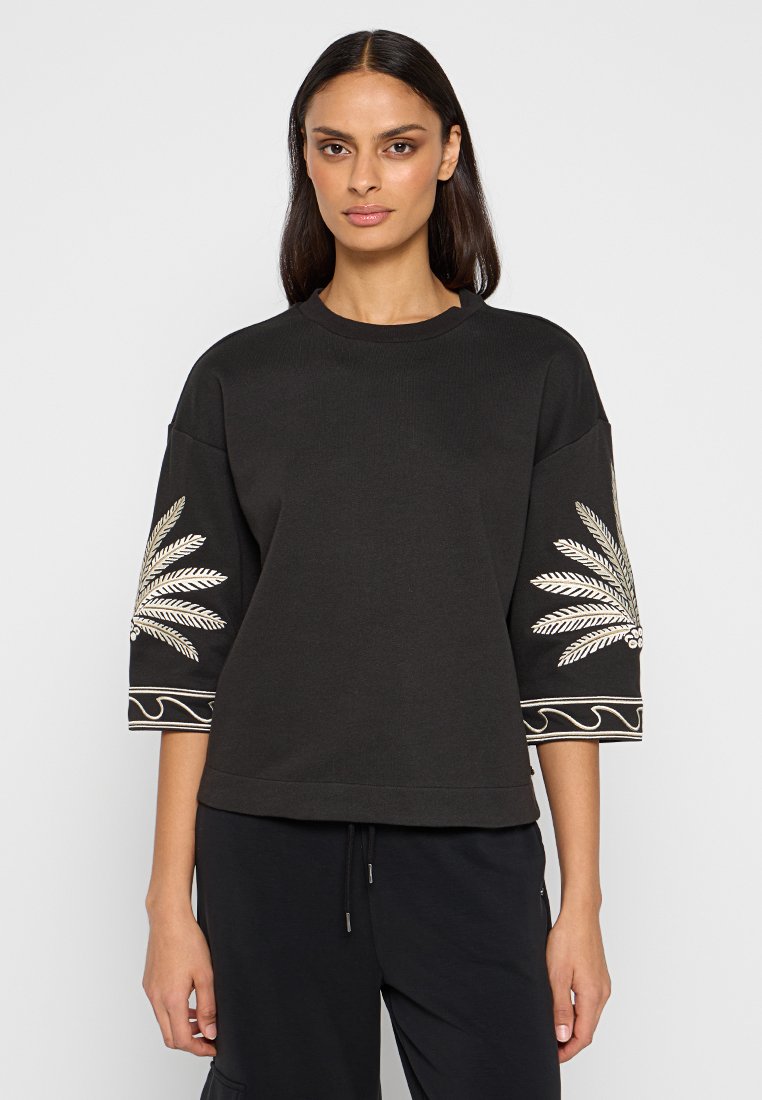 Sudadera negra de manga corta, con un diseño bordado de hoja de palma en blanco y acentos dorados en las mangas. Detalle de dobladillo elástico.