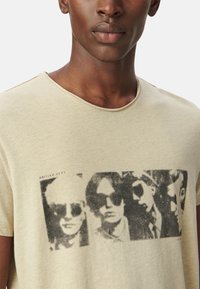 Camiseta de algodón beige con cuello redondo, que presenta una impresión gráfica negra de rostros con gafas de sol. Corte casual, mangas cortas.