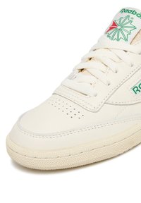 Krémovo biele tenisky Reebok spredu s bielymi šnúrkami, perforovanou špičkou a zeleno-červeným logom na jazyku na bielom pozadí.