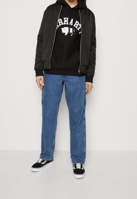 Giacca bomber nera sopra un hoodie nero con logo bianco, abbinata a jeans larghi blu e sneakers nere con suole bianche.