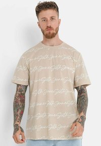 Beige bomull t-skjorte med gjentatt hvitt håndskrevet tekstmønster, rund hals og korte ermer. Tatoveringer synlige på armene.