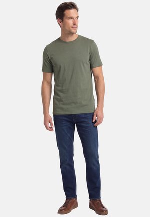 Man draagt een olijfgroen T-shirt met korte mouwen, donkerblauwe spijkerbroek en bruine leren sneakers, staat met ontspannen handen en kijkt naar rechts.