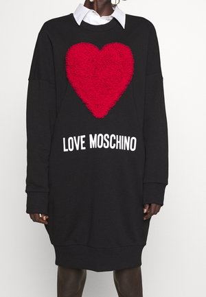 Czarna sukienka z długim rękawem, na przodzie ozdobiona dużym, fakturowanym czerwonym sercem oraz napisem "LOVE MOSCHINO", noszona na białej koszuli z kołnierzykiem.