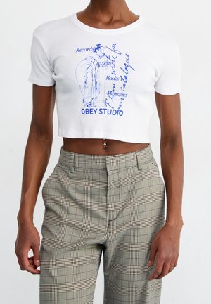 Osoba ubrana w biały krótki top z niebieskim tekstem i ilustracją, w połączeniu z beżowymi spodniami w kratę o wysokim stanie na jednolitym tle.