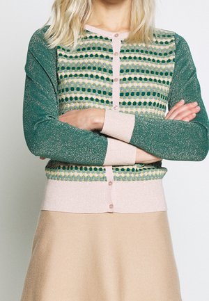 Femme aux cheveux blonds portant un pull boutonné vert à motifs avec des poignets beiges et une jupe beige, les bras croisés.