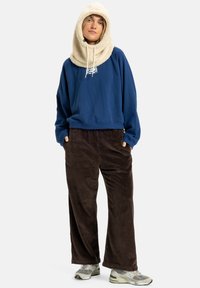 Modrá mikina s logem, krémová kapucí šála, hnědé kordové kalhoty a šedé tenisky. Pohodlný a ležérní outfit s vrstvenými texturami.