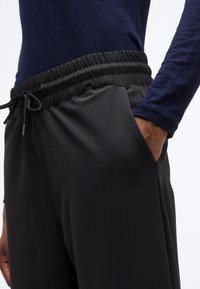 Άτομο που φορά μαύρο παντελόνι με ελαστική μέση και κορδόνι, και μπλε σκούρο πουκάμισο με μακριά μανίκια, με το χέρι στην τσέπη.